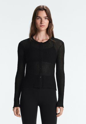 Junge Frau mit langen braunen Haaren, die eine figurbetonte schwarze, durchsichtige gerippte Strickjacke mit Knöpfen und schwarze Hose trägt, steht vor einem einfarbigen hellen Hintergrund.