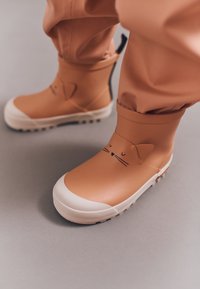 Bottes imperméables beiges avec des semelles blanches, présentant un design de visage de chat avec des oreilles et des moustaches. Texture lisse avec un ajustement flexible.