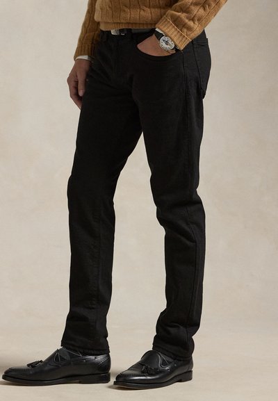 Férfi fekete nadrágban, fekete bojtos mokaszinban, barna kötött pulóverben és karórával, egy kézzel a zsebében áll.