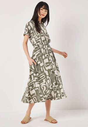 Mujer con vestido midi de patrón abstracto verde y blanco, con mangas cortas y cintura elástica, posando con una mano en el bolsillo y sandalias beige.