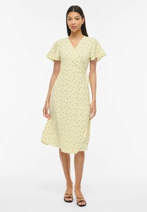 Femeie purtând o rochie galbenă cu flori, tip înfășurare, cu mâneci scurte evazate și sandale negre cu vârfuri deschise, stând pe un fundal alb.