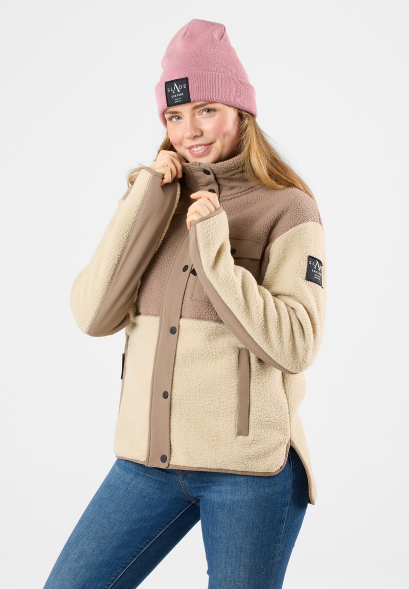 Giacca in pile beige e marrone con chiusura a zip e bottoni a pressione, dotata di due tasche frontali e colletto imbottito. Indossata con un berretto rosa.