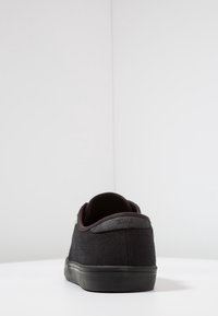 Musta kangaskenkä, jossa on teksturoitu pinta, pyöreä kärki ja kontrastivärinen kumipohja. Varustettu logolla kantapäässä ja yksinkertaisella designilla.