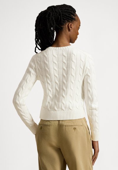 Beli pulover z označbo kabelskih pletenin z okroglim izrezom, robom z rebrastim vzorcem in dolgimi rokavi, kombiniran z krojenimi kaki hlačami z zadrgami na zadnjih žepih.