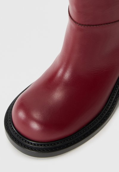 Burgundy leather boot with a smooth finish, rounded toe, and a black textured sole featuring decorative stitching along the edge.