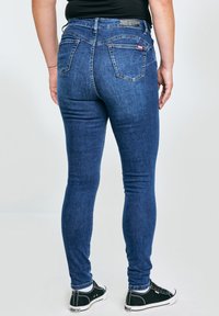 Osoba u uskim plavim trapericama i crnim tenisicama stoji okrenuta leđima prema kameri, na jednobojnoj bijeloj pozadini.