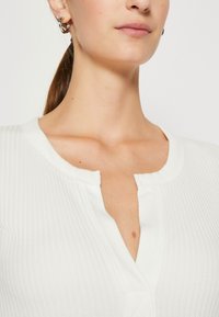 Vit ribbad stickad topp med rundad krage och knäppning vid halsen. Slät textur och rena linjer är tydliga.