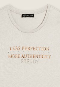 T-shirt corto color beige chiaro con scollo rotondo. Presenta testo in rame metallico e strass, con scritto "MENO PERFEZIONE PIU’ AUTENTICITÀ."