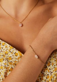 Gouden ketting en armband met roze stenen accent, gezet tegen een bloemrijke gele achtergrond. Textuur van de huid zichtbaar.