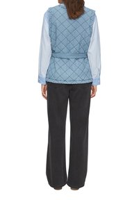 Gep quiltete hellblaue Weste mit einem taillierten Gürtel über einem blau-weißen gestreiften Hemd. Schwarze weit geschnittene Hosen runden das Outfit ab.