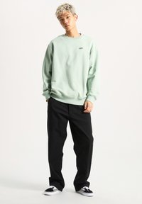 Sudadera verde clara con cuello redondo, corte relajado y pequeño logo. Pantalones negros con diseño de pierna recta, combinados con zapatillas de cuadros en blanco y negro.