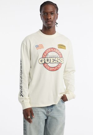 Crème lange mouwen shirt met het "GUESS" logo, Amerikaanse vlag en patchaccenten. Zachte katoenstof met een ontspannen pasvorm. Lichte jeans.