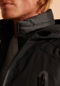 Chaqueta negra acolchada con un cuello gris texturizado, cierre de cremallera y detalle de logotipo. La tela es lisa con un acabado mate.