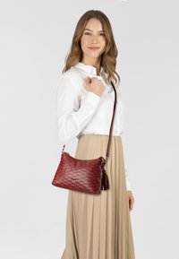 Bolso de mano de cuero rojo con un patrón acolchado, herrajes plateados y un acento de borlas. Forma compacta, llevado con una correa cruzada.