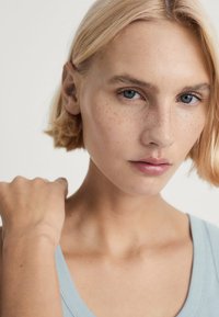 Detailný záber na osobu so svetlo blond vlasmi a modrými očami, oblečenú v svetlomodrom tielku s hladkým materiálom a voľným strihom.