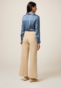 Camicia di satin azzurro chiaro con dettaglio sul retro, abbinata a pantaloni beige a gamba larga con tasche posteriori e un taglio pulito e sartoriale.