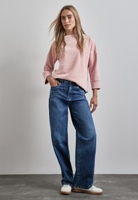 Helles rosa geripptes Strickoberteil mit lockerer Passform, weite blaue Denim-Jeans und weiße Sneaker vor einem neutralen Hintergrund. Lässiges Outfit präsentiert.