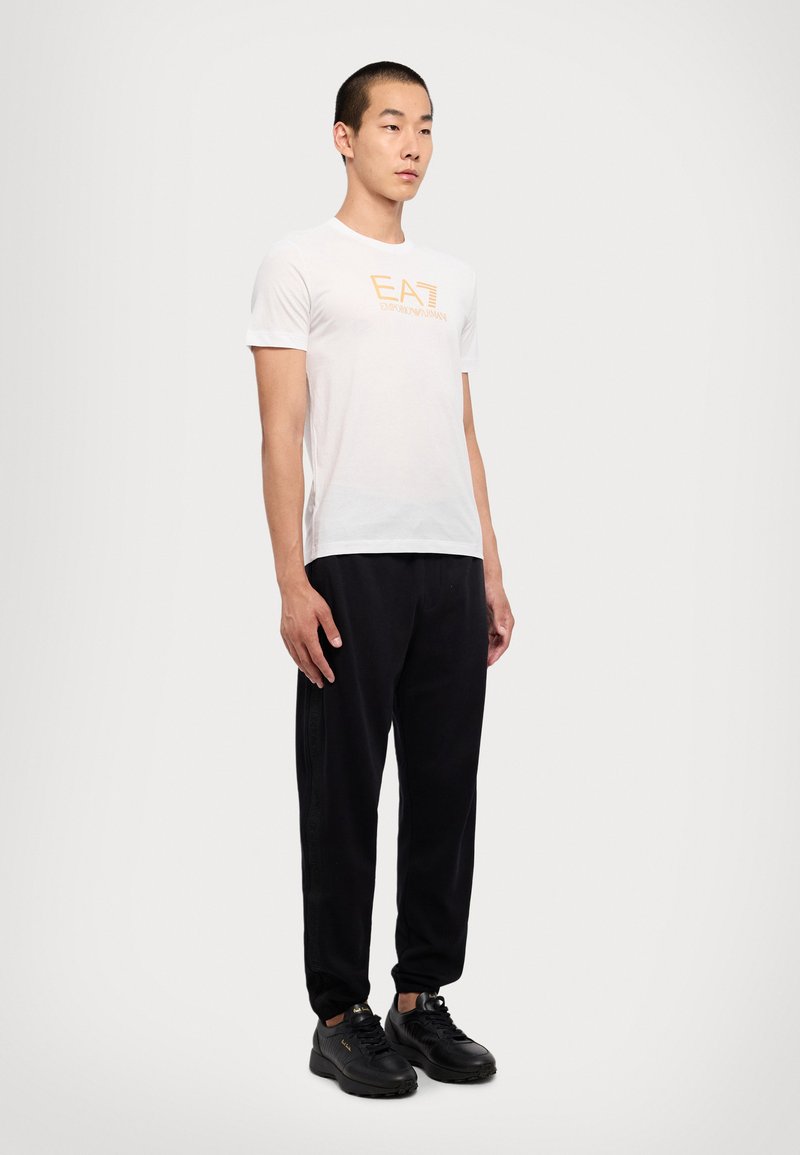 Hvitt crew-neck t-skjorte med oransje logo, svarte tapered joggebukser og svarte sneakers. Enkel design, glatt tekstur og casual passform.
