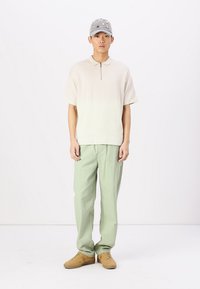 Polo beige chiaro a maniche corte con zip, abbinato a pantaloni verde chiaro e sneakers color cammello. Cappellino grigio con decorazioni.