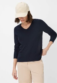 Marineblauer ribbeliger Wollpullover mit V-Ausschnitt und langen Ärmeln, kombiniert mit beigen Hosen und einer hellen Mütze. Schlichtes, tailliertes Design.