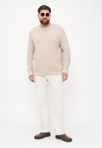 Beige texturovaný svetr s kulatým výstřihem, doplněný bílými kalhotami a hnědými botami. Modelka nosí kulaté černé sluneční brýle.