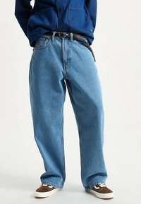 Jeans di denim blu a gamba larga con vita alta, cuciture visibili e cintura nera. Abbinati a sneakers marroni con lacci bianchi.