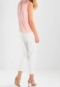 Blouse rose pâle sans manches avec encolure froncée, associée à un pantalon blanc ajusté et des talons à bride beige. Texture de tissu lisse.