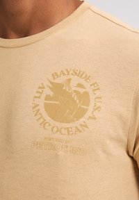 Beige katoenen t-shirt met een ronde halslijn en korte mouwen, voorzien van een vervaagde grafische print van een vis en tekst.