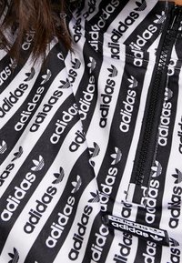 Černobílá bunda na zip s opakujícím se vzorem loga „adidas“, hladkou texturou a polyesterovým materiálem. Viditelný výrazný brandingový detail.