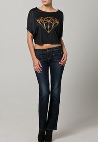 Černý zkrácený top se zlatým geometrickým diamantovým designem, spárovaný s tmavými denimovými džíny s pěti kapsami a rozšířenými nohavicemi.