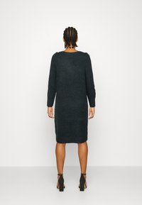 Abito a maglia verde scuro, lungo fino al ginocchio, con maniche lunghe e scollatura rotonda. Tessuto liscio, spacchi laterali, indossato con sandali neri con tacco alto.