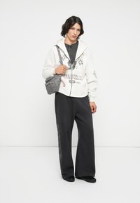 Felpa bianca con zip e stampa grafica, t-shirt grigia a trama sotto, pantaloni neri larghi, scarpe nere slip-on e una piccola borsa a tracolla grigia.