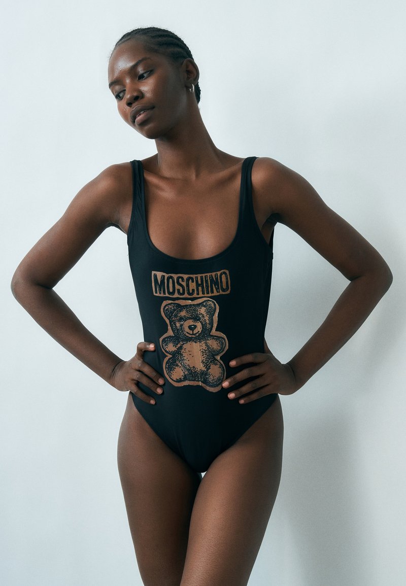 Γυναίκα με πλεγμένα μαλλιά που φοράει μαύρο μαγιό με μεγάλο αρκουδάκι και το κείμενο "Moschino", στέκεται με τα χέρια στη λεκάνη.