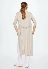 Long cardigan ouvert en beige, avec des manches longues, un col châle et des poches latérales. Le tissu paraît doux et tombe gracieusement.
