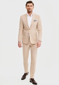 Beżowy garnitur z lekkiego materiału, składający się z marynarki z klapami i slim-fit spodni. Połączony z białą koszulą i ciemno-brązowymi butami.