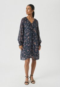 Vrouw in een marineblauwe bloemenjurk met lange mouwen en ruches, met zwarte enkellaarsjes met enkelband, staand en naar de zijkant kijkend.