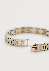 Brățară metalică cu tonuri alternante de argintiu și auriu, cu finisaj texturat, închidere cu cataramă și un mic logo dreptunghiular în culorile roșu, alb și albastru.