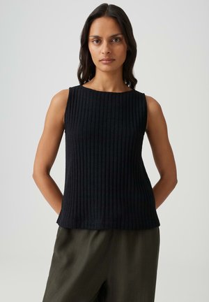 Femme aux longs cheveux foncés portant un haut noir côtelé sans manches et un pantalon vert olive, debout devant un fond clair uni.