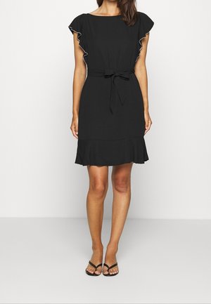 Woman wearing a knee-length black dress with flutter sleeves, a waist tie, and ruffled hem, paired with black open-toe sandals.