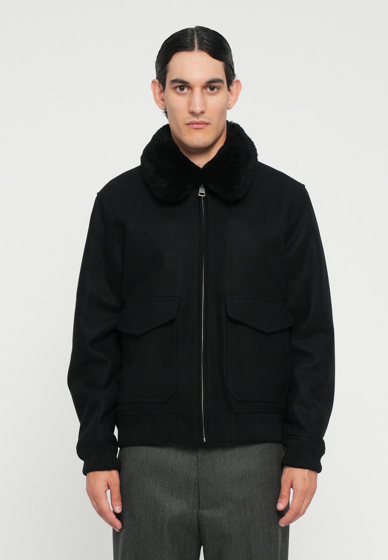 Schwarze Bomberjacke mit einem plüschigen schwarzen Kragen, Frontreißverschluss und zwei aufgesetzten Taschen. Rippbündchen und -saum, strukturierte Passform, glatte Textur.