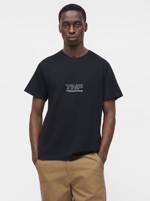 Mand iført en sort T-shirt med "TNF The North Face" logo og beige bukser, stående med hænderne i lommerne mod en ensfarvet baggrund.