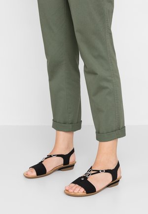 Black suede sandals with a T-strap design, circular silver accents, and a low heel, paired with rolled-up green pants.