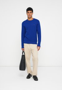 Kék textúrált kötött pulóver, kerek nyakkivágással, bézs nadrággal és fekete loaferekkel párosítva, fekete tote táskával a kezében.