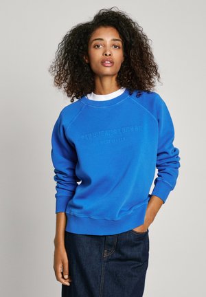 Sudadera azul con puños y dobladillo acanalados, con texto de logotipo en relieve. Emparejada con una falda de mezclilla oscura, el modelo muestra un atuendo informal.