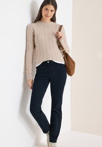 Femme portant un pull beige à torsades, un pantalon bleu foncé et des baskets blanches, portant un sac à bandoulière marron, appuyée contre un mur.