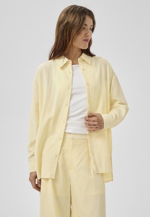 Naine kannab lahtist helerohelist nööpidega särki valge topi kohal ning sobivaid helerohelisi laia säärega pükse, vaadates vasakule.