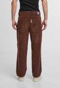 Brown corduroy pants with a straight-leg design, featuring patch pockets at the back and a branded tag at the waistband.