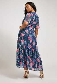 Abito maxi floreale in un profondo blu navy con motivi di fiori rosa e blu. Caratterizzato da maniche trasparenti e orlo a balze a strati. Indossato con sandali con tacco dorati.