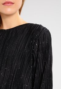 Svart plisserad topp med texturerad tyg och glittrande detaljer, rund halsringning och långa ärmar. Fokusera på vertikala plisseringar och glans.