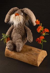 Lapin en peluche marron avec des oreilles tombantes et une barbe blanche, tenant une carotte en tissu, assis sur une bûche en bois à côté de fleurs orange sur un fond sombre.